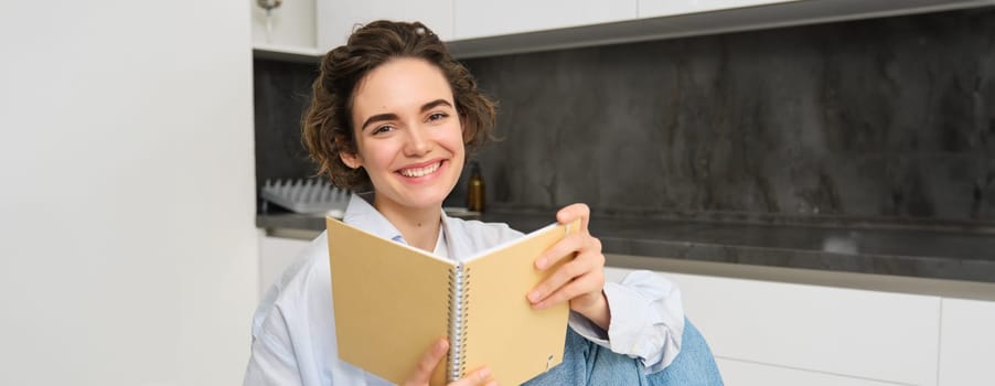 Image of beautiful girl at home, holding notebook, doing her homework, studying for exam in her kitchen and smiling.