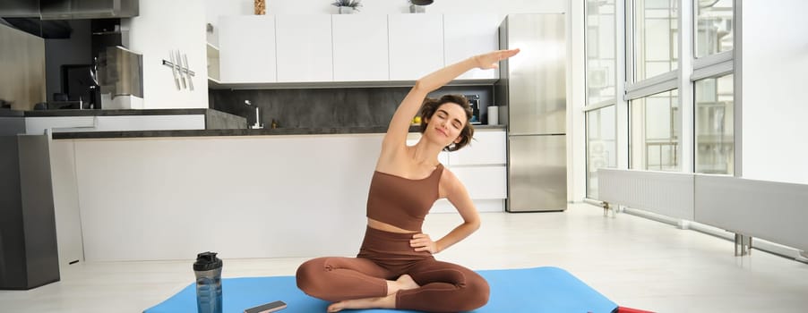 Portrait of fitness woman doing exercises in bright room at home, stretching in lotus pose, sitting on rubber yoga mat and doing workout training, drinking water from bottle.