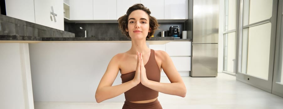 Close up portrait of young woman at home, sitting and meditating, practice yoga, making lotus pose, exercise indoors. Sport and women wellbeing concept