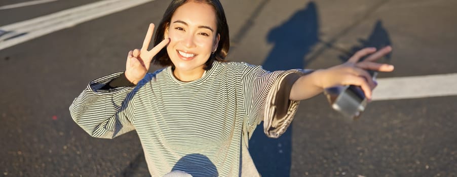 Selfie of asian girl sitting on skateboard, taking photo on smartphone, smiling and showing peace v-sign.