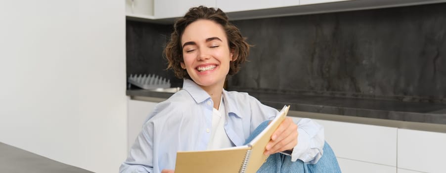Portrait of happy smiling woman, holding notebook, studying in her kitchen, reading journal. Lifestyle and people concept