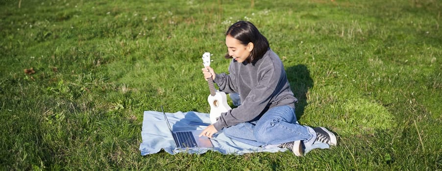 Happy smiling asian girl enjoying spring time, playing ukulele in park, looking for chords online on laptop, learning new song.