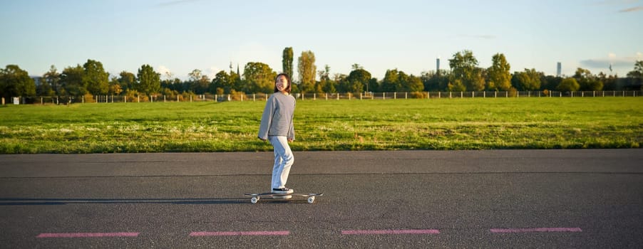 Beautiful asian skater girl riding her longboard on sunny empty road. Young woman enjoying her skate ride smiling and laughing.