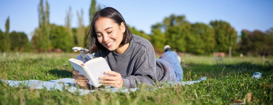 Portrait of beautiful smiling asian girl, reading in park, lying on grass with favourite book. Leisure and people concept.