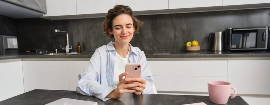 Portrait of beautiful smiling woman sitting at home with smartphone, using mobile phone app, orders groceries delivery.