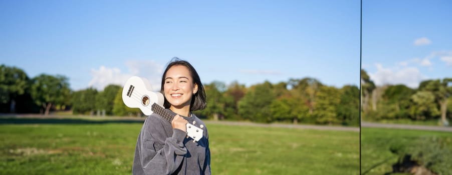 Young hipster girl, traveler holding her ukulele, playing outdoors in park and smiling. People concept