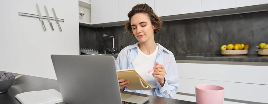 Working woman makes notes, writes down information on notebook, sitting with laptop at home.