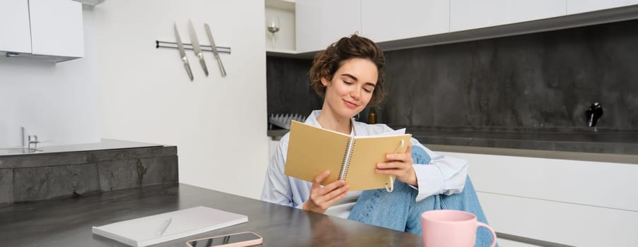 Smiling beautiful woman, sitting with notebook in kitchen, reading notes, studying, doing homework.