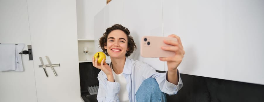 Beautiful girl blogger, takes selfie as she eats apple, shows how to lead healthy lifestyle to followers online, holds smartphone for photo.