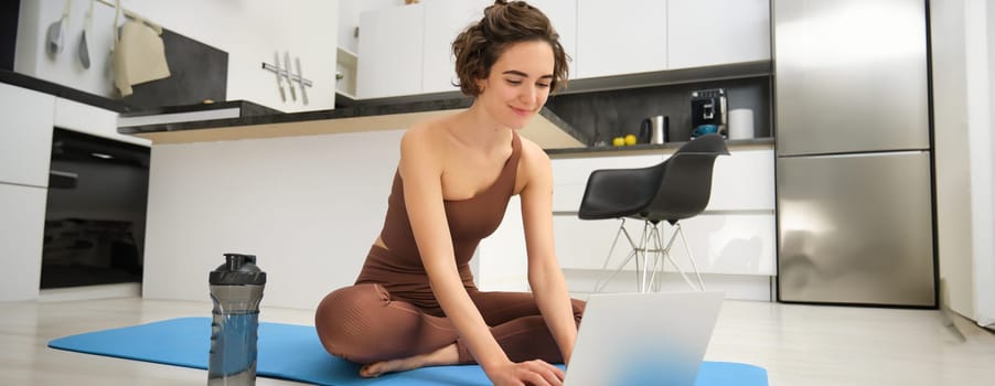 Portrait of young female athlete, fitness instructor starting online gym class from her home, sitting on rubber yoga mat, typing on laptop. Sport and lifestyle concept