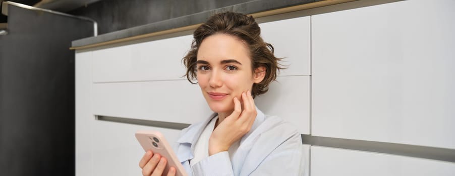 Portrait of girl with mobile phone, sits on floor at home, orders takeaway in app. Young woman with smartphone does online shopping from home, browsing websites.