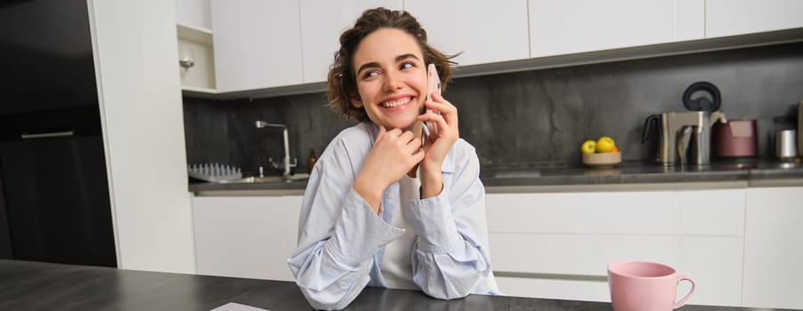 Smiling young woman talks on mobile phone, calls someone from home, sits in kitchen and has conversation. Copy space