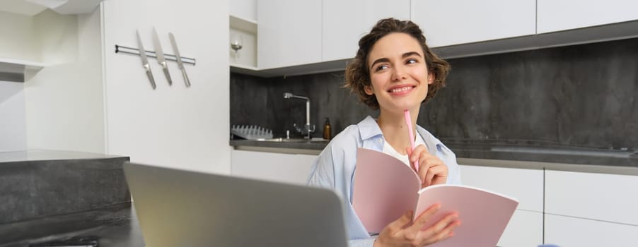 Young smiling woman works from home, writes in her diary, makes notes in journal planner, sits near laptop in kitchen.