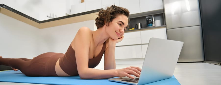 Portrait of woman athlete, girl workout at home, watches fitness videos on laptop, lying on rubber mat and doing indoor yoga session. Sport and wellbeing concept