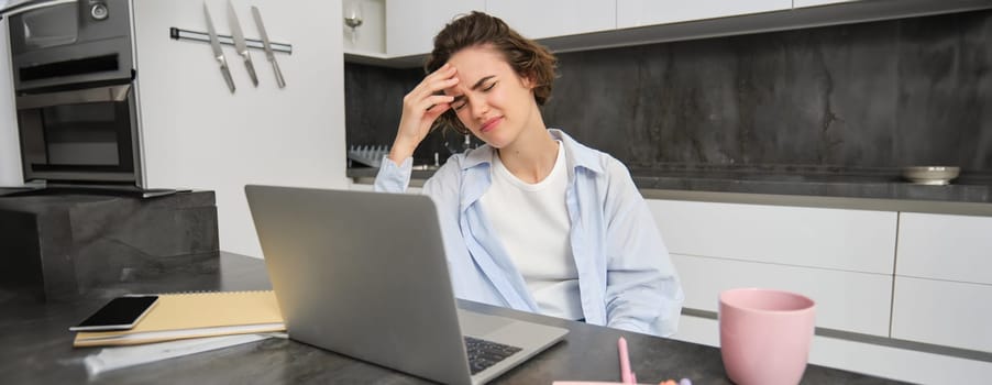 Woman has headache while works from home. Girl has painful migraine, sits near laptop, grimaces from discomfort.