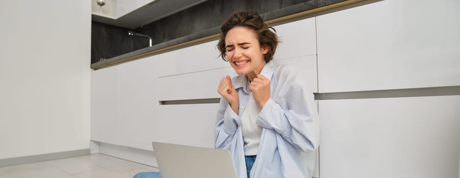 Candid, smiling young woman sits on floor at home, works remotely, connects to online group chat meeting, using laptop to talk with friend.