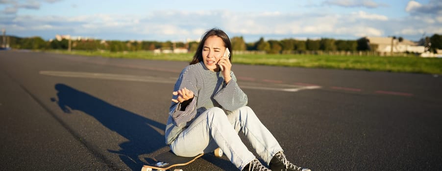 Portrait of asian girl sits on her longboard, skating on skateboard and talking on mobile phone, having conflict during telephone conversation, arguing with concerned face.