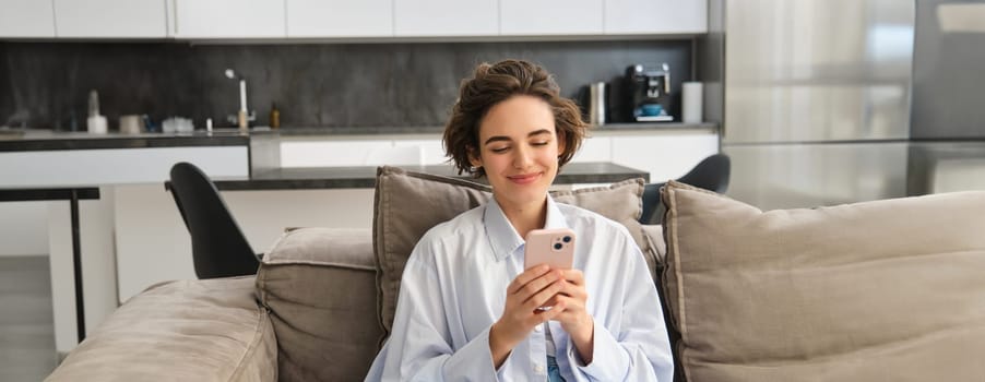 Image of happy woman sits on couch with smartphone, looks at mobile phone screen with pleased smile, chats in app, watches videos online.