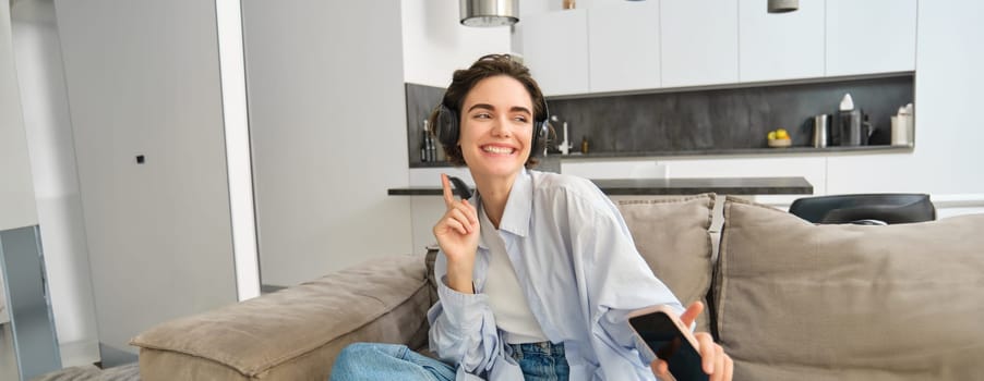 Portrait of happy girl in headphones, dancing on sofa and listening music, having fun at her house.