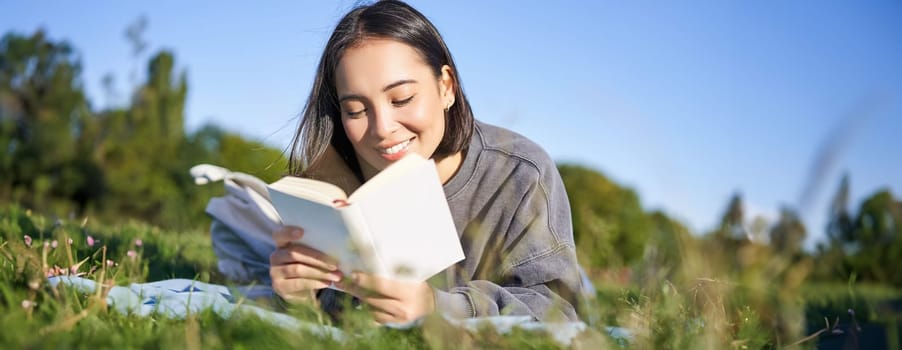 Portrait of cute korean girl, reading in park while lying on grass, relaxing with favorite book in hands, smiling happily.