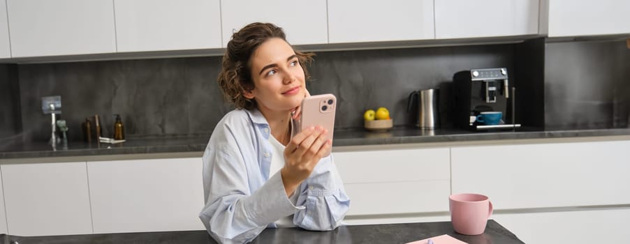 Technology and lifestyle. Young woman sits at home, uses smartphone in her kitchen and smiles.