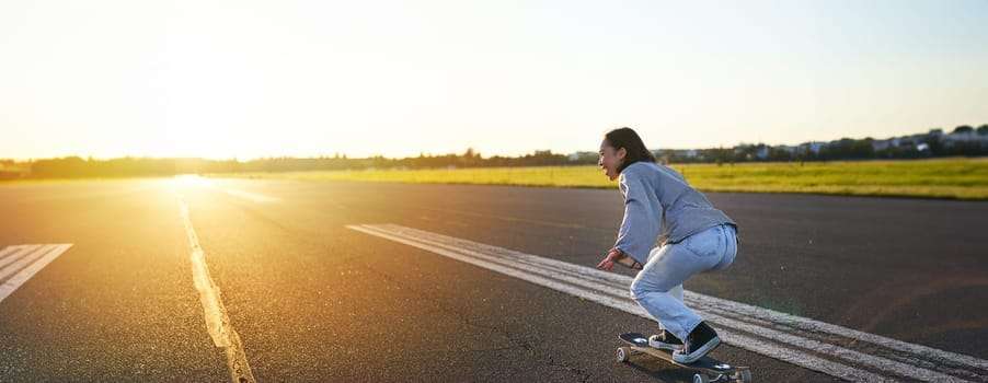 Side view of beautiful asian girl on skateboard, riding her cruiser towards the sun on an empty road. Happy young skater enjoying sunny day on her skate.