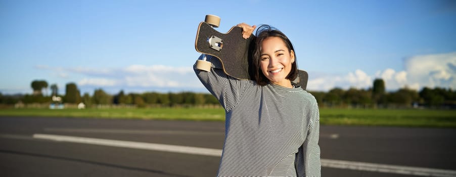 Portrait of beautiful young skater girl, standing with longboard and smiling at camera. Asian woman with skateboard standing on road.