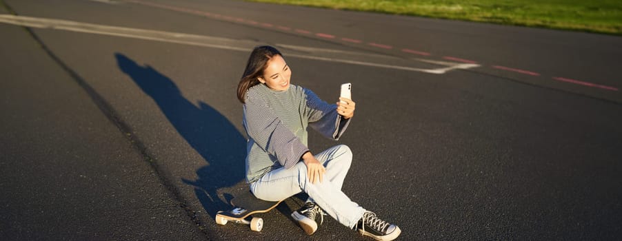 Beautiful korean girl takes selfie on smartphone, takes photo with her skateboard, enoys sunny day outdoors.