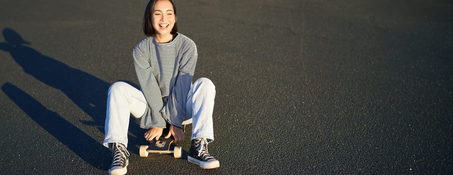 Freedom and happiness. Cute smiling asian girl, sits on skateboard on sunny spring day. Happy laughing skater enjoying cruising on longboard.