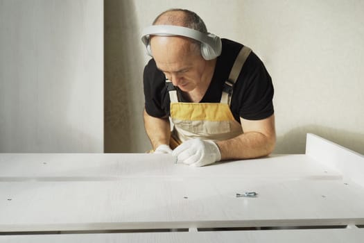 Furniture assembly. A worker fastens the parts of a bed using a confirmat. While working, he listens to music on headphones. Close-up.