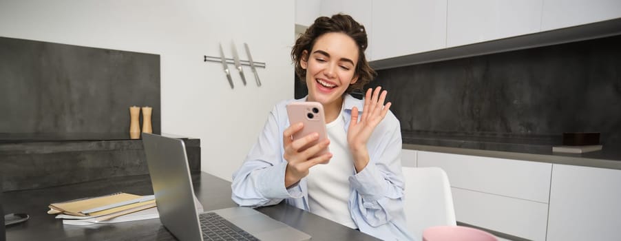 Portrait of young woman video chats with friend on smartphone, waves hand at mobile phone camera, calls someone using application.