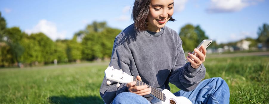 Technology and music. Smiling asian hipster girl sitting in park with smartphone and holding ukulele, playing instrument.