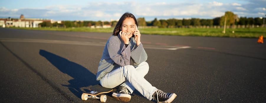 Cute teen girl sits on skateboard and talks on mobile phone. Happy skater woman having conversation on smartphone.