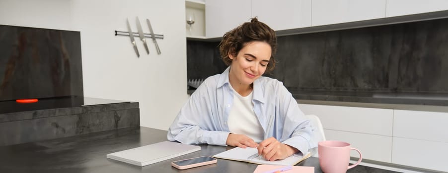 Portrait of young woman working from home, writing down information in notebook, taking notes, sitting in kitchen and studying, student doing homework.