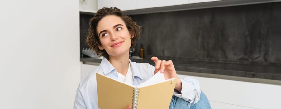 Portrait of young adult woman, reading her notes, holding notebook and smiling, relaxing at home.