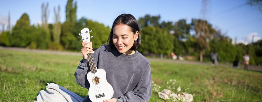 Vertical shot of happy asian girl plays instrument, shows her ukulele at laptop camera, video chats about music, teaches how to play, sits in park outdoors.