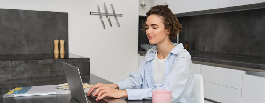 Working woman using laptop, drinking coffee. Girl with cup of tea, studying from hyome, sitting near computer in kitchen, preparing for exam.