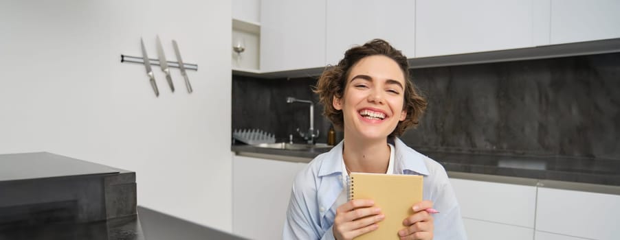 Portrait of young creative woman, holding her notebook and smiling, makes notes in her diary, writing in her planner, sitting at home in kitchen and smiling.