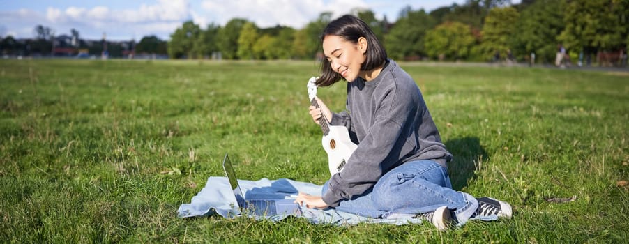 Smiling asian girl learns how to play ukulele via laptop, online video tutorials, sitting on grass in park with musical instrument.