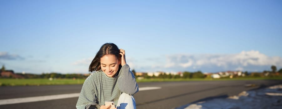 Vertical shot of asian woman sitting on skateboard on road, holding smartphone app. Skater girl skates on longboard, using mobile phone.