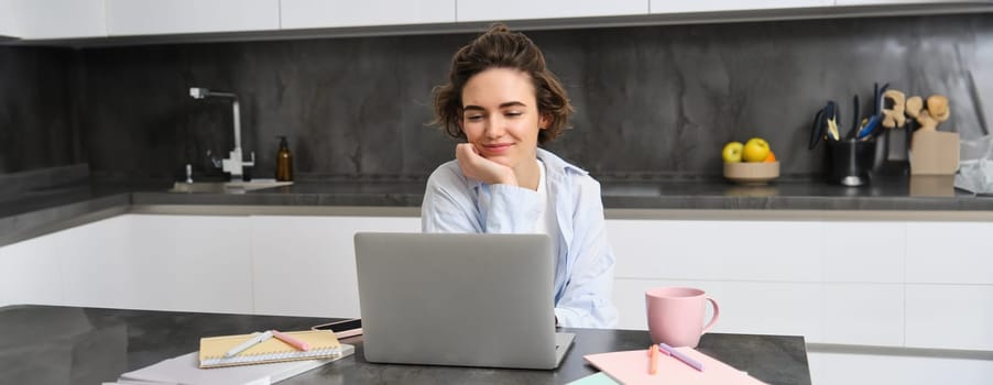 Portrait of young professional woman, works from home, looks at laptop, studies online, connects to a webinar, work remote meeting via computer, video chats with tutor from courses website.