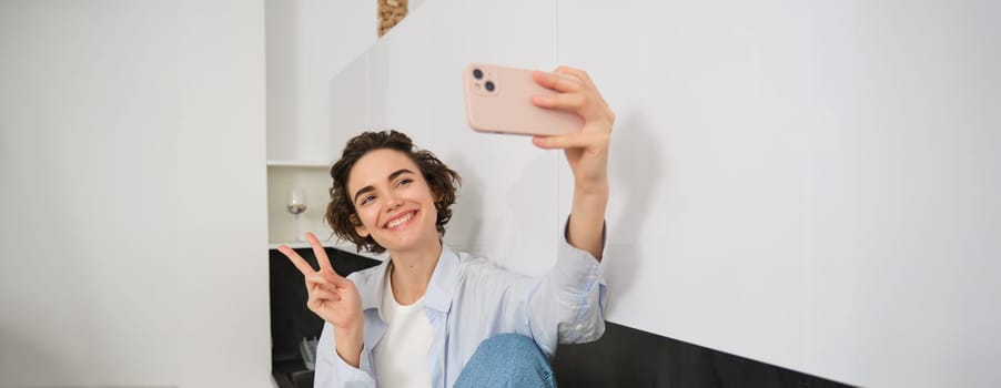 Portrait of beautiful brunette girl, takes selfie on smartphone in her kitchen, posing for photo on mobile phone.