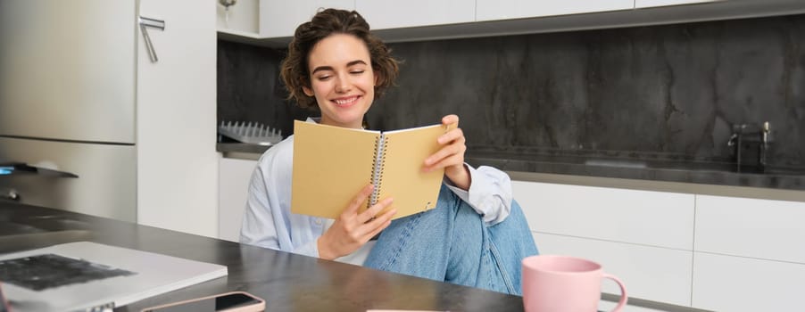 Smiling beautiful woman, sitting with notebook in kitchen, reading notes, studying, doing homework.