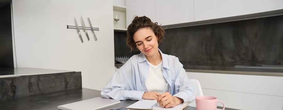 Portrait of young woman working from home, writing down information in notebook, taking notes, sitting in kitchen and studying, student doing homework.