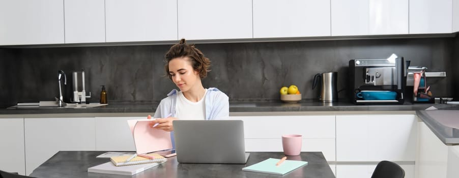 Portrait of working woman at home, checks her notebook, flips page, connects to online meeting via laptop. Girl learns programming remotely, studies course in internet.