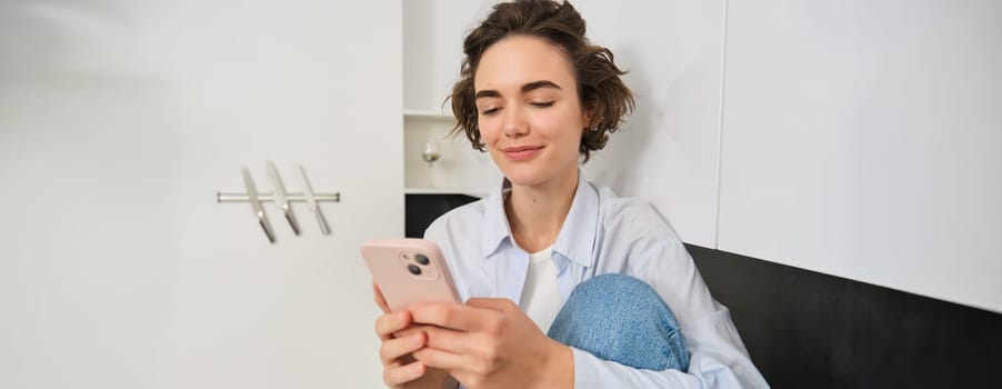 Portrait of young woman looking at mobile phone, reading a message, watching smth on smartphone app at home, smiling while looking at screen.