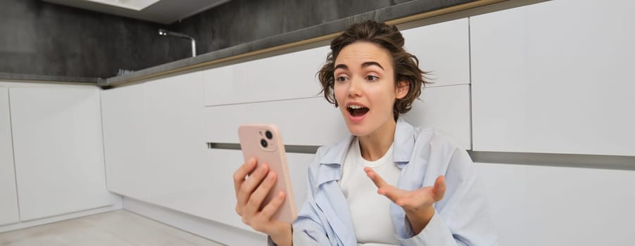 Portrait of amazed, candid young woman, holding smartphone, reacts surprised at big news, video chats with someone, sits on kitchen floor.