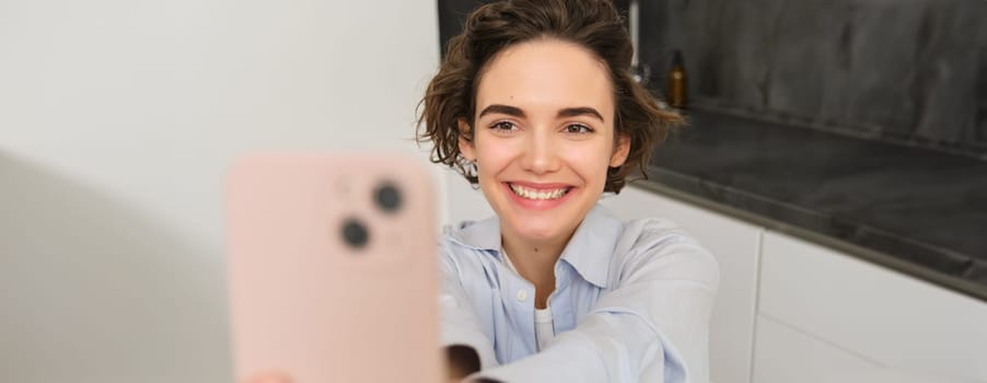 Portrait of beautiful young woman, taking selfie on mobile phone at home, poses for photo with smartphone.