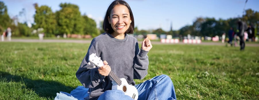 Portrait of excited asian girl, sitting with ukulele in park on green grass, looking amazed and surprised with good news.