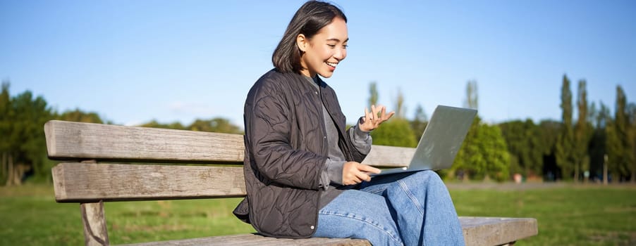 Portrait of young asian woman working remotely from park, sitting on bench with laptop, talking during video chat.